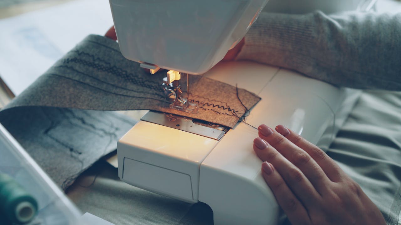 hero-img-01 Close-up of a sewing machine stitching fabric, focused on hands and textile.
